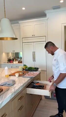 A man wearing a chef's jacket is in a modern kitchen. He is pulling open a drawer in the kitchen island. The island has a marble countertop. On the countertop there is a cutting board, a bottle of olive oil, a grey cloth, a small cup with green contents, a tray of raw chicken, a bunch of parsley, and a bottle of yellow liquid. Behind the island, there is a refrigerator and white cabinets. To the left of the refrigerator, there is a doorway leading to a dining area. The dining area has a round table set for a meal. Beyond the dining area, there is a living room with a television, a bookshelf, and seating areas. A white floor fan is positioned near the doorway.