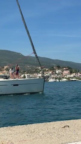 The video captures a bright, sunny day at a harbor in Poros, Greece. A large, modern white sailboat with a furled dark sail is seen slowly traversing calm blue waters. Two individuals are on the boat's deck: one person, wearing a white top and light-colored bottoms, is bent over near the bow, appearing to handle lines or equipment. The second person, in darker attire, stands upright further back on the deck.

From an alternate perspective, a person is shown standing on a concrete pier in the foreground, holding a white shopping bag. This individual wears a navy and white striped long-sleeved shirt and white shorts. The background features rolling green hills dotted with a cluster of buildings, characteristic of a coastal Greek town, under a clear blue sky. The scene depicts a leisurely moment, possibly during a boat's arrival or departure from the harbor.