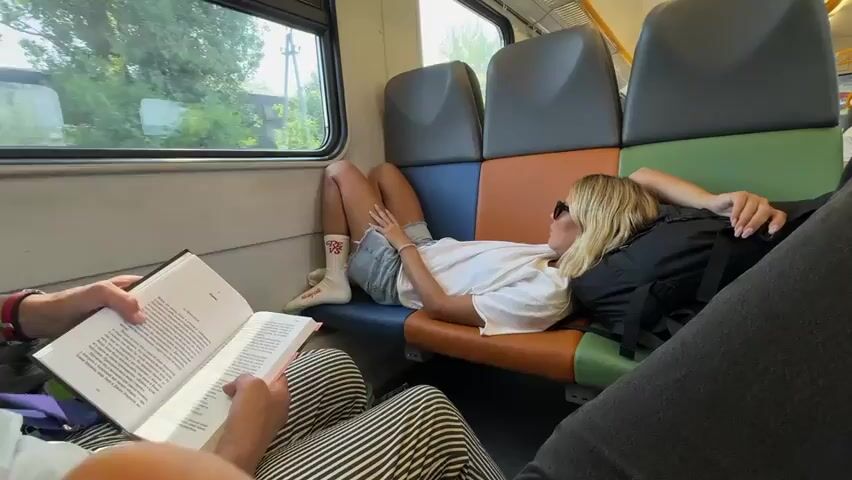 The video shows the interior of a moving train car. The camera is positioned to show two passengers. In the foreground, a person is reading a book with a white cover. Their arms and a portion of their striped pants are visible. In the middle ground, a blonde woman is lying down on the train seats, with her head resting on a black backpack. She is wearing a white t-shirt, denim shorts, and white socks with red accents. Her legs are extended and her arm is resting on her stomach. She is looking out the window. Behind her, the seats of the train are visible, with different colored sections: blue, orange, gray, and green. Through the window, trees and greenery can be seen, indicating the train is traveling through a rural or suburban area. The sunlight is bright, casting shadows inside the train car. Another passenger, whose shoulder and arm are visible, is seated to the right of the woman. The overall atmosphere suggests a relaxed and casual journey.
