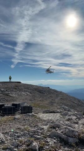 A helicopter hovers in the air above a rocky, mountainous landscape. The sky is blue with white, wispy clouds, and the sun is visible in the upper right corner. In the foreground, there are several large, dark rectangular objects arranged on the ground, possibly crates or storage containers. To the left, a person wearing a yellow safety vest stands on a platform overlooking the scene. The helicopter appears to be either landing or taking off, as it is positioned at a low altitude. The overall environment suggests a remote, possibly construction or research site in a high-altitude location.
