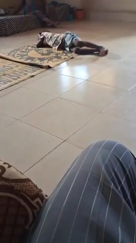 The video depicts a room with tiled floors, a woman sitting on a bed, and a child lying prone on a rug. The woman is seated towards the back of the room, wearing a patterned cloth. The child is lying face down on a rug, with their body stretched out. A second rug is partially visible to the left of the child. The room is lit by natural light coming through a window, casting shadows on the floor. A person is seated in the foreground, with their patterned cushion and striped trousers partially visible. The person in the foreground seems to be filming the scene. An orange pot sits on the bed near the woman.