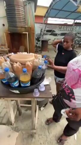 The video shows a woman purchasing what appears to be a juice drink from a roadside vendor. The vendor is standing behind a small wooden table laden with plastic bottles of various colored liquids, arranged in a circular display within a black plastic tub or tray. There are at least 10-15 bottles visible, some with blue caps and others with brown caps.  Some bottles have labels on them.  The woman selects a bottle and takes it from the vendor. In the background, another younger woman is standing near a building with a “CARWASH” sign. The setting appears to be an outdoor commercial or industrial area, with a large metal tank visible behind the vendor's table.  The vendor hands the woman the bottle of juice.