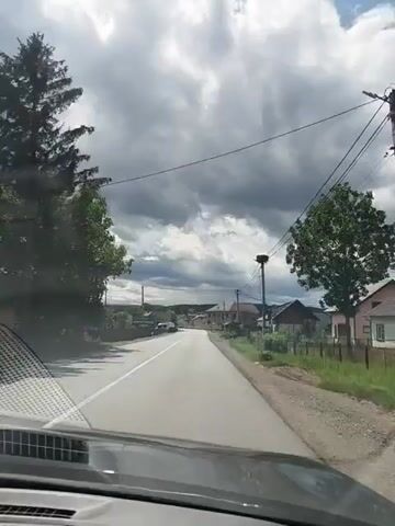 The video shows a point-of-view perspective from inside a vehicle driving on a paved road. The road is lined with trees on the left and houses and fences on the right. The sky is filled with dark, dramatic clouds, suggesting impending rain. There are power lines running along the right side of the road, supported by utility poles. In the distance, there are more houses and buildings. A few vehicles are visible on the road, including a white van and a car. The video captures the motion of the vehicle as it travels through what appears to be a rural or suburban area.