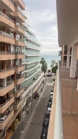 The video, taken from a balcony, overlooks a street leading to a beach. On either side, tall hotel buildings with glass-railed balconies rise. Several people can be seen on the balconies and strolling down the street. The street has parked cars, a crosswalk, and a few palm trees. A red "STOP" sign is visible at the corner. Beyond the street, the turquoise sea stretches to the horizon under a cloudy sky. A small beach with a lifeguard hut is visible at the end of the street.