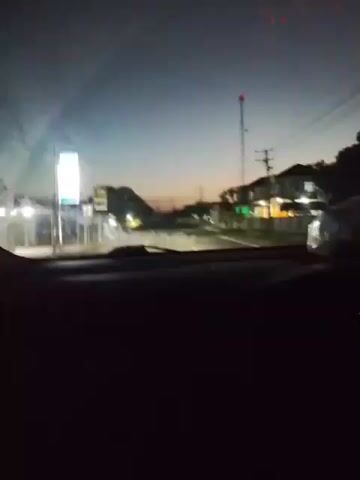 This is a first-person perspective video of a vehicle moving at night. The view is from inside the car, looking out through the windshield. The sky is dark blue, suggesting it's either dusk or dawn. The road ahead is dimly lit, with some buildings and trees visible on either side. There are streetlights and lights from the buildings, creating a soft glow. Electrical poles and wires are also visible. The dashboard of the car is partially visible in the lower part of the frame, showing some illuminated dials. The footage is somewhat blurry and shaky, indicating movement.