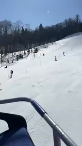 The video shows a skier riding a ski lift up a snowy slope on a sunny day. Other skiers can be seen skiing down the hill. The landscape is a snow-covered mountain with trees. The sky is blue with some clouds. The video is taken from the perspective of the skier on the lift, looking down the slope. The lift structure and support pillars are visible in the frame.