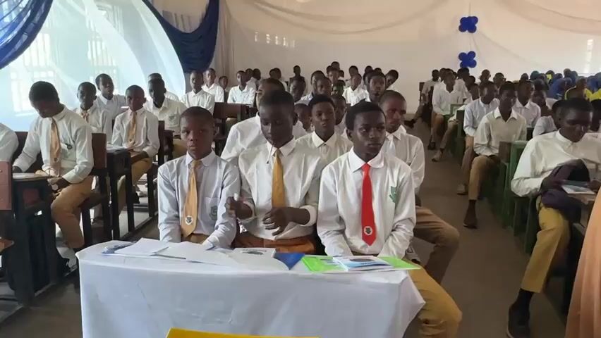 The video captures a large gathering of students, mostly young males and some females, seated in rows within what appears to be a school hall or event space in Unguwan Dosa, Nigeria. The students are dressed in school uniforms consisting of white long-sleeved shirts, ties of various colors (yellow, red, green), and khaki or brown trousers. Several female students are wearing hijabs in shades of brown and blue.

In the foreground, three male students are seated behind a white-draped table, with papers and books visible on the table surface. The student in the middle wears a yellow tie, and the one on the right wears a red tie. The rest of the students fill the background, seated on chairs arranged in orderly rows.

The room is decorated with white drapes along the ceiling and walls, accented with blue fabric drapes and clusters of blue and white balloons, suggesting a formal event or celebration. The students appear to be attentive, with most looking towards the front of the room, presumably at a speaker who is not visible in the frame. A male voice can be heard speaking, indicating an ongoing presentation or address. The overall atmosphere suggests an educational or motivational event.