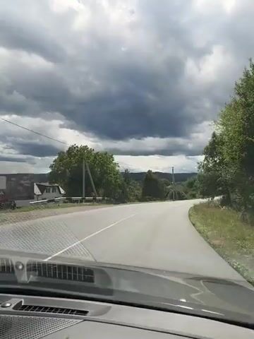 The video shows a view from inside a car driving on a paved road. The road curves to the right. There are trees and vegetation on both sides of the road. In the distance, there are hills and a cloudy sky. There are power lines visible overhead. On the right side of the road, a house is visible. The car appears to be moving at a moderate speed.