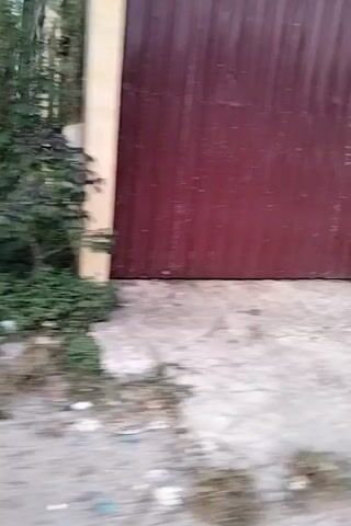 The video shows a ground-level view of an outdoor scene. A red corrugated metal shutter is visible on the left side of the frame, with a tan-colored wooden beam above it. A small, reddish-brown box-like object is attached to a rough, light-colored wall on the right. As the camera pans, a partially constructed building with exposed wooden scaffolding and brickwork comes into view. A motorcycle is parked in front of a covered area with wooden supports and a corrugated roof. Further along, there's a glimpse of some greenery and a muddy or wet patch on the ground. The video appears to be shot at an angle and may be shaky, indicating movement during filming. The environment seems to be a somewhat neglected or under-construction area, possibly in a rural or semi-urban setting.