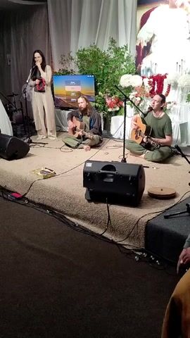 A woman in a light-colored outfit stands at a microphone, holding a tambourine, while two men sit on the floor playing guitars. A large speaker is positioned on the stage, with cables running across the floor. A television screen displays a landscape image. The stage is carpeted, and in the background, there are curtains, plants, and floral arrangements. A large picture or tapestry hangs on the wall behind the performers. The overall setting appears to be an indoor performance space, possibly a spiritual or cultural event.