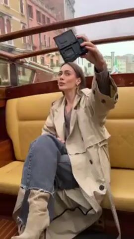 A woman sits in a boat, holding up a small black rectangular object, manipulating it, and taking a photo with it. She is wearing a beige trench coat, jeans, and heeled boots. The boat has a tan leather interior. Outside the boat are buildings, suggesting she is in Venice. The overall lighting suggests an overcast day.