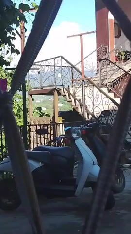 The video pans across a scene viewed through what appears to be a metal grate or fence. In the foreground, two scooters are parked. The scooter on the left is white and grey, and the scooter on the right is dark. Behind the scooters, a set of outdoor stairs with ornate metal railings ascends to a building. Lush green foliage is visible to the left of the stairs, and the sky above is blue with scattered clouds. The overall impression is an outdoor setting, possibly a courtyard or residential area.