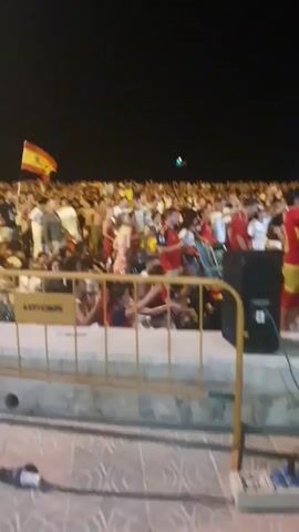 The video shows a large crowd of people gathered at night. The crowd appears to be watching or participating in an event, as many are looking in the same direction. Some individuals are holding flags, one of which is red and yellow, possibly the Spanish flag. People are standing, sitting, and some appear to be dancing. There are barriers in the foreground, suggesting a controlled viewing area. The overall atmosphere seems to be one of excitement and celebration. The background is dark, indicating nighttime, with a few lights visible in the distance. A yellow sign with text "AVTY CALPE" is visible on a barrier in the foreground.