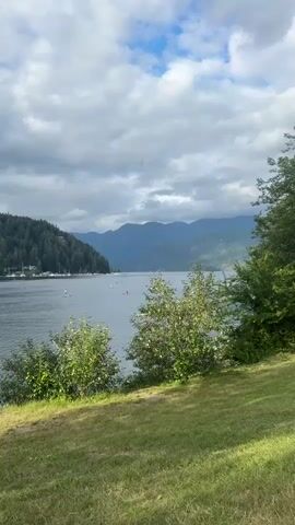 A wide shot shows a serene body of water, likely a lake or bay, with calm ripples. The water is bordered by lush green hills covered in dense evergreen trees on the left and a more distant range of majestic, misty blue mountains in the background. The sky above is a mixture of white, puffy clouds and darker, overcast gray clouds, suggesting a partly cloudy day. In the foreground, a grassy slope slopes down towards the water, with several bushes and trees providing a natural frame. Scattered across the water are various recreational watercraft, including kayaks, canoes, and what appear to be paddleboards. People are visible in some of these boats, engaged in activities like paddling and observing the scenery. A few sailboats are also docked or sailing near the shore on the left side of the frame. The overall atmosphere is peaceful and invites a sense of outdoor recreation and natural beauty.