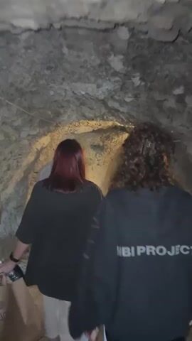 Three individuals are seen walking away from the camera through a narrow, dimly lit, rocky tunnel or cave. The tunnel walls and ceiling are composed of rough, light-colored rock with some areas illuminated by artificial yellowish light. The floor is a dirt path. The person closest to the camera has curly brown hair, is wearing dark sunglasses and a black hoodie with "MBI PROJECT" printed in white letters on the back. This person briefly glances back at the camera. In front of them, a person with red hair is wearing a black t-shirt and white pants, carrying a brown paper bag and a clear plastic bottle in their left hand. A third individual, wearing a light-colored top, is visible further ahead, leading the group.