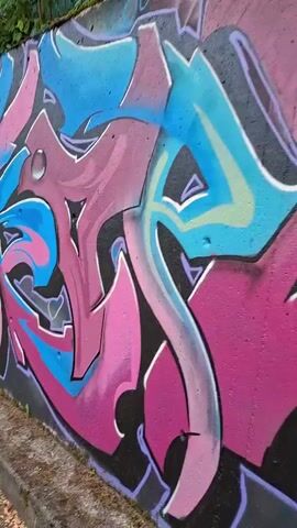 The video shows a mural painted on a concrete wall. The mural features a stylized owl's face with large, bright yellow eyes and a blue beak. The owl is surrounded by abstract graffiti art in various colors, including pink, blue, and purple. The graffiti has a sharp, angular style with bubble-like accents.

The scene is outdoors, with a concrete curb and asphalt road visible at the bottom of the frame. Some greenery, including grass and weeds, grows along the curb and the base of the wall. A green metal fence is visible at the top of the wall, suggesting the wall might be part of a boundary or overlook.

The graffiti and the owl art are the primary subjects. The activity is the display of street art. The time of day appears to be daytime, based on the lighting. The weather seems to be clear. The location context provided is Forbach, Germany. There is no visible text in the video, other than what is incorporated into the graffiti design itself, which is not clearly decipherable. The overall impression is one of vibrant and dynamic urban art.