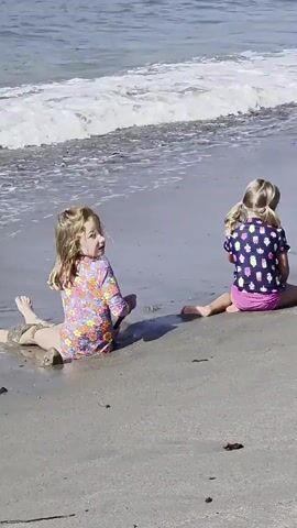 Two young girls are sitting on a sandy beach near the ocean. The girl on the left is wearing a colorful long-sleeved rashguard with a floral pattern and is leaning forward, her hands in the wet sand near the water's edge. Her hair is styled in braids. The girl on the right is wearing a dark t-shirt with a pattern of colorful popsicles and pink shorts. She is sitting with her back to the camera, facing the ocean. Both girls appear to be enjoying a day at the beach. The waves are gently rolling in, and the water looks clear. The sky is bright, suggesting it is daytime, likely mid-morning or afternoon, with sunny weather. The scene is typical of a beach outing, with the children focused on their activities in the sand and by the water. There is no visible text in the media.