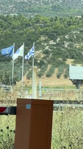 A video shows a border crossing with flags of the European Union and Greece flying. There is a barrier gate and a small building in the background. The landscape is hilly and covered with trees. The video is shaky and appears to be filmed from a moving vehicle.