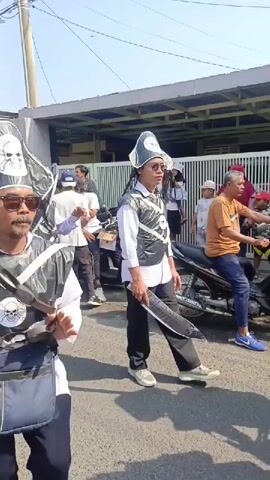 A group of men in pirate-themed costumes are walking down a street. They are wearing hats adorned with skull and crossbones symbols and vests made of black plastic. Some of the men are carrying what appear to be fake swords or scimitars. They are walking in a procession, with some participants interacting with each other and others looking towards the camera. The environment appears to be an outdoor street scene with buildings and a fence visible in the background. There are also motorcycles and other people present, suggesting a public event or parade.