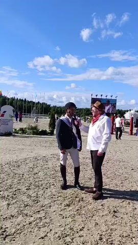 This video captures an outdoor scene on a sunny day with scattered clouds, suggesting it's midday. The setting appears to be an equestrian event, likely a horse show or competition, given the sandy ground, fencing, and distant spectators and structures associated with such events.

In the foreground, two individuals are the primary focus. On the left stands a man dressed in riding attire, including a helmet, dark jacket, white breeches, and tall black boots. He wears a sash that appears to be for an award or recognition, possibly indicating he is a participant or winner. He is holding a microphone and seems to be engaged in an interview.

To his right, another man, dressed in a more casual yet festive manner, is speaking into a microphone, interviewing the rider. He wears a brown hat, a white shirt, a red scarf around his neck, and dark pants. His posture and interaction suggest a reporter or commentator.

In the background, several other people are visible, some walking and others gathered near tents or structures. Flags are planted in the ground along a fence line, and a large screen is mounted on a structure, possibly for displaying event information or replays. The overall atmosphere appears to be relaxed and celebratory.

While no specific text is clearly legible on clothing or signs, the presence of the sash and the interviewing setup strongly suggest an awards ceremony or a post-competition interview at an equestrian event. The bright sunlight and blue sky indicate pleasant weather conditions. The sandy surface is characteristic of riding arenas.