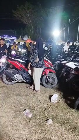 The video shows a nighttime outdoor gathering with many people and parked motorcycles. The lighting is artificial, coming from various sources, including spotlights and lights on tents in the background. The ground appears to be dirt or grass, and there is some litter visible. In the foreground, several motorcycles are parked closely together. People are seen standing near the motorcycles, some sitting on them, and others walking around. The individuals in the video are dressed in casual clothing, with some wearing headscarves. The overall atmosphere appears to be that of a public event or festival.