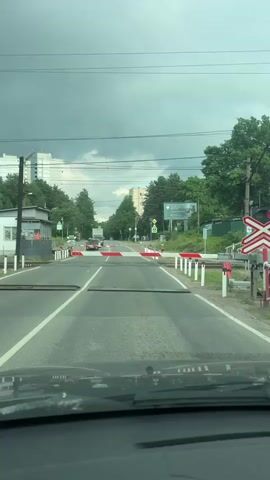 The video shows a view from inside a vehicle approaching a railroad crossing. The crossing gates are down, indicated by red and white striped barriers. There are also metal plates across the road, possibly to prevent vehicles from crossing the tracks when the gates are down. A red traffic light is visible on a pole to the right, also indicating that vehicles should stop. A few cars are stopped at the crossing, and a truck is visible in the distance. The sky is cloudy. Trees line the sides of the road, and buildings can be seen in the background.