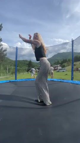 A woman with blonde hair and black-and-white striped pants is jumping on a trampoline. The trampoline has a blue padded rim and a net enclosure. In the background, there is a grassy field with a few people standing around and a mountain range under a partly cloudy sky. The woman successfully executes a back tuck on the trampoline.