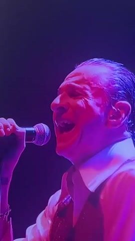 A close-up, side profile shot shows a man singing into a microphone. He is wearing a white collared shirt and a vest. The lighting is dramatic, with blues and purples creating a moody atmosphere. The man's hair is slicked back, and he appears to be performing on stage.