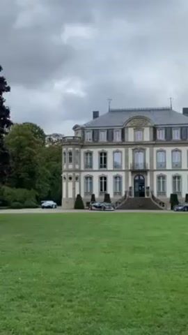 The camera pans upward on a cloudy day, showing a stately three-story manor house with a gray slate roof, light beige siding, and ornate stone trim. A sprawling green lawn extends to the viewer, with several luxury cars parked near the house. Trees border the sides of the frame, suggesting the property is surrounded by woods.