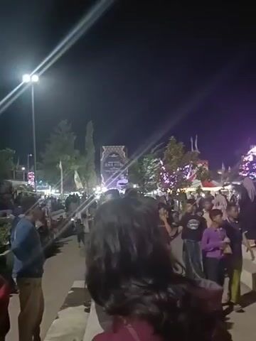 The video shows a nighttime outdoor scene filled with a large crowd of people. They appear to be gathered in a public area with tiered seating, similar to bleachers or stadium steps. There are streetlights illuminating the area, as well as decorative lighting in the background near an archway-like structure. The people are mainly standing or sitting, and there is a sense of anticipation or excitement in the air. Some are looking in a specific direction, possibly towards a stage or focal point. Several individuals in the foreground are partially visible, including a young girl in a maroon jacket. Overall, it gives the impression of a public event or gathering taking place at night.