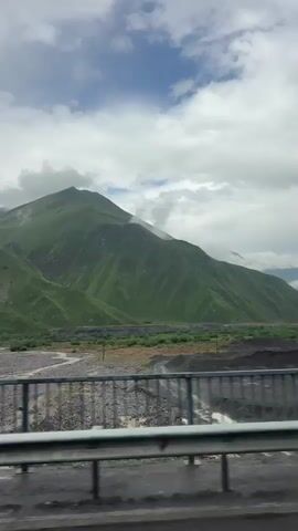 The video shows a wide shot of a mountainous landscape with a riverbed in the foreground. The mountains are covered in lush green vegetation, and the sky is cloudy with patches of blue visible. A metal railing runs across the bottom of the frame, suggesting the viewer is on a bridge or elevated roadway. The riverbed appears to be rocky and dry, with some signs of recent water flow. In the distance, there are industrial structures and piles of dark material, possibly related to mining or construction. The camera is stationary, capturing the scene from a fixed viewpoint. The lighting is natural, suggesting it is daytime.