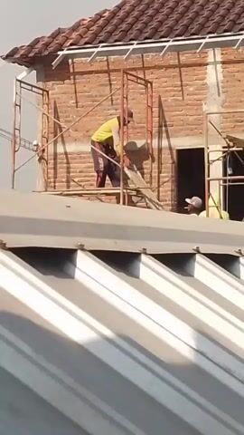 The video shows construction workers on a building. One worker in a yellow shirt and red pants is standing on scaffolding and holding a wooden plank. Another worker in a light green shirt is lying on a roof below, and a third worker, also in a light green garment, is sitting near the second worker. The building is made of brick, and scaffolding is erected around it. The roof is covered with terracotta tiles. The scene appears to be during the daytime, with natural light illuminating the area.