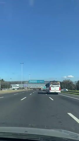 The video shows a car driving on a highway under a sign that reads "Bienvenidos a la Provincia" which translates to "Welcome to the Province". A white bus is in front of the car. A toll plaza is further ahead. The sky is blue.