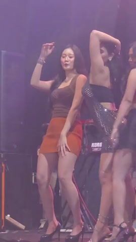The video shows three women dancing on a stage. The woman on the left wears a brown top and an orange skirt. The woman in the middle wears a sparkly black dress. The woman on the right wears a red top and a black skirt. They are all wearing high heels and are moving their bodies to the music. The stage is lit with purple and green lights. There are speakers and other equipment visible on the stage.