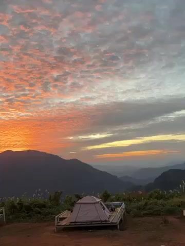 The video shows a scenic view of a mountain range at sunrise or sunset. The sky is filled with textured clouds, colored with shades of orange, pink, and grey. In the foreground, there are some raised wooden platforms, with a tent set up on one of them. A man appears to be taking a photo with his phone. A woman then walks into frame. There is a white plastic chair on the ground.
