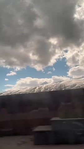 The video shows a view from a moving vehicle, possibly a car or train. In the foreground, there's a blurry, dark-colored coniferous tree passing by. Behind the tree, a low, dark-colored wall or fence runs horizontally. The background features a distant hill or mountain range covered with patches of snow. Above the snowy hills, the sky is visible, showing a mix of blue and thick, greyish-white clouds, creating a partly cloudy atmosphere. The overall scene seems to depict a landscape viewed from a traveling vehicle on a partly cloudy day.