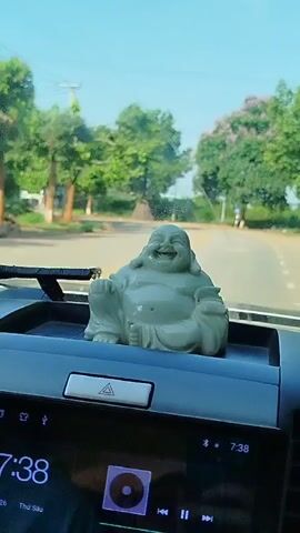 From inside a vehicle driving along a paved road, a small gray Buddha statue sits on the dashboard above a touch screen display showing "7:38". The camera is still while the car is in motion. Green trees line the road outside the vehicle. A small structure is visible on the right side of the road.