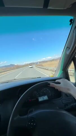 A person is driving a car on a highway on a clear, sunny day. The view is from inside the car, focusing on the steering wheel and the road ahead.  The driver has a visible hand with black nail polish. The landscape consists of a paved road with mountains in the distance.  A road sign is visible, with distances to Chita and Londonka. Other cars are on the road. The sky is bright blue with some clouds.