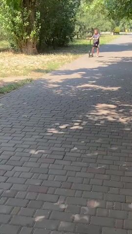 A boy rides a scooter on a paved path. The path is made of grey bricks and has shadows cast on it from trees. There are trees and grass on either side of the path. The boy is wearing a purple shirt and black shorts and has a bag slung over his shoulder. He is riding away from the camera.