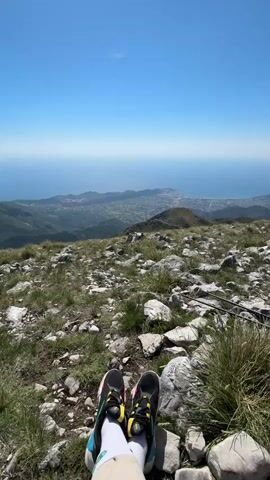 The video shows a person relaxing on a rocky hillside, with their feet and lower legs in view. They are wearing black and yellow sneakers with white socks and khaki pants. The ground is covered in rocks and patches of green grass. In the distance, there is a view of a coastline and a city. The sky is clear and blue. A voice can be heard in the background. The video pans slightly to the left, showing more of the rocky terrain.
