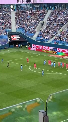 A football match is underway in a stadium packed with spectators. Two teams, one in red and the other in blue, are on the field. The red team is lined up in a defensive wall, anticipating a free kick from the blue team. The ball is visible near the penalty area, and the blue team players are positioned to take the shot. The stadium is brightly lit, and advertising boards are visible around the pitch. A person is visible in the foreground, looking out over the field.