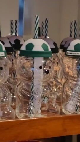 The video shows a row of clear, bear-shaped drinking cups on a wooden shelf. Each cup has a green and white soccer ball-shaped lid with a green band that says "STARBUCKS". A green and white striped straw goes through the lid of each cup. The cups are transparent, revealing a white paper insert inside, which has Starbucks branding on it. In the background, there are more similar cups, some with slightly different colored soccer ball patterns. The lighting appears to be indoor, and the cups are arranged neatly in a display.