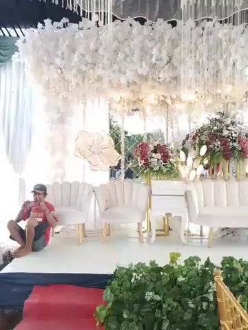 The video shows a wedding venue. The stage is decorated with white floral arrangements hanging from the ceiling. There are also floral arrangements of red and white flowers on pedestals on the stage. White furniture is arranged in the center of the stage including two chairs and a love seat. A man is sitting on the floor taking pictures of the stage with his phone. The stage is in front of a mirror. Greenery and gold chairs line the front of the stage. A red carpet is in front of the stage.
