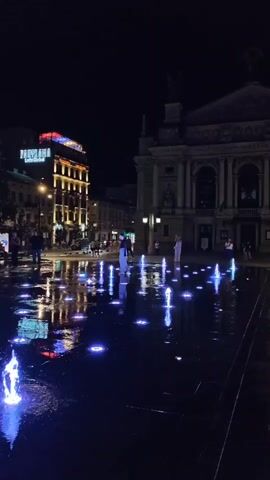 A night time shot of a public square featuring a water fountain with blue and pink lights. Several people are walking around the fountain, some are standing and watching the lights. In the background, a large ornate building, possibly a theater or opera house, is visible, as well as a modern building with illuminated signs. The ground around the fountain is wet and reflects the lights. The overall atmosphere is lively and urban.