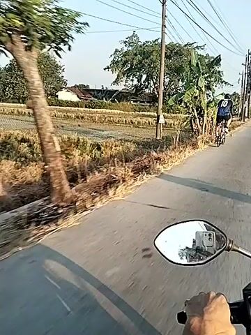 A first-person perspective video shows a person riding a motorbike on a paved road. In front of the motorbike, a cyclist wearing a black jersey with the number 10 on the back is also riding on the road. The cyclist is ahead of the motorbike and appears to be moving in the same direction. The environment is rural, with fields on the side of the road, banana trees, and utility poles with wires. The sun is shining, casting shadows. The video cuts out briefly showing a close-up of the cyclist from a different angle, and then resumes showing the original perspective.