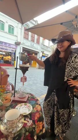 A woman wearing a cowboy hat and a black outfit with a patterned overlay pours liquid from a kettle into a coffee dripper set up on a table. The table is covered with a floral tablecloth and has a teacup and saucer on it. In the background, people walk by on a cobblestone street in front of buildings. The scene appears to be outdoors, under an awning.