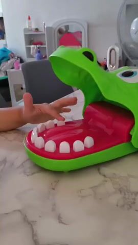 The video shows a close-up of a child's hands playing with a green crocodile toy. The toy has a large open mouth with red interior and white teeth. The child's hands are positioned inside the mouth of the toy, suggesting they are interacting with its mechanism. The background appears to be an indoor setting, possibly a home, with a marble-patterned tabletop. A blurred background shows a shelf unit with various items, a chair, and a mirror, indicating a domestic environment. The lighting suggests it is daytime. No weather or specific time of day information is available, beyond it being daylight. There is no visible text in the media file.