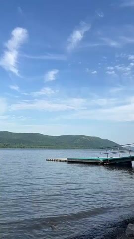 The video begins with a view of a body of water, with mountains in the background. A pier extends into the water with a ramp leading down to a lower section. The camera then tilts upwards to capture the sky, which is a bright blue with scattered white clouds. A small airplane flies across the sky, leaving a contrail behind it.