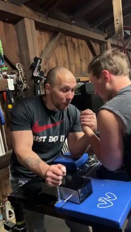 Two men are arm wrestling in what appears to be a garage. The man on the left wears a black t-shirt with a red Nike logo and the words "Just Do It," jeans, and has tattoos on his left arm. The man on the right wears a grey tank top. Both men are leaning over a blue arm wrestling table, gripping each other's hands. A black metal box with a lever is situated between them on the table. Various tools and equipment hang on the wooden walls in the background. The man on the left is speaking and demonstrating how to position his thumb to prevent slipping.