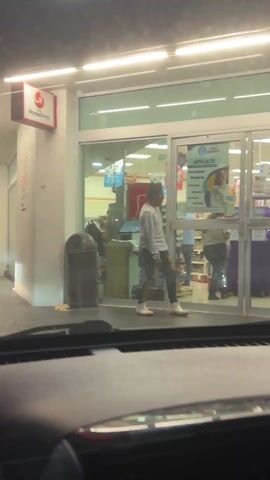 A man exits a Super Farmacia store. He is wearing a light gray sweater, patterned pants that are primarily dark with yellow streaks, and white boots. The store's interior is visible through the glass doors and windows, including shelves stocked with products, a cash register area, and other customers. Signage for the pharmacy is displayed on the door, advertising affiliation and listing medications. A trash can is positioned outside the door to the left. The video is shot from inside a vehicle.